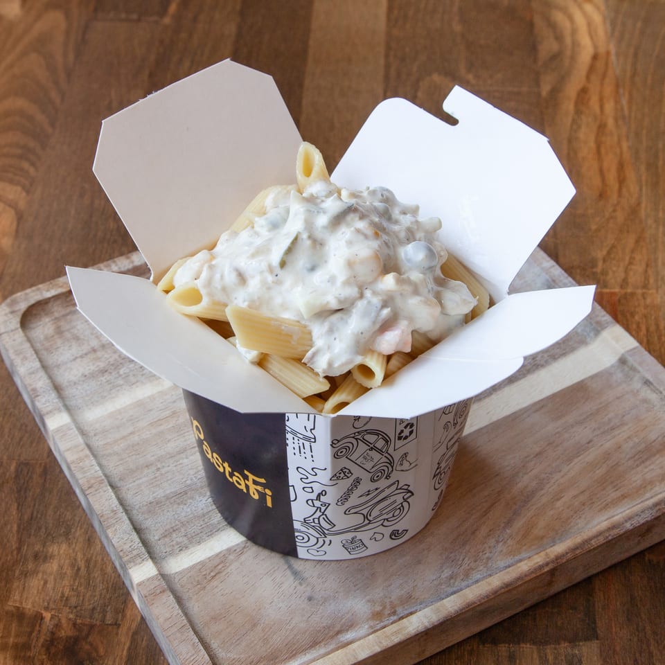 Takeout container filled with penne pasta topped with creamy white sauce on a wooden serving board.