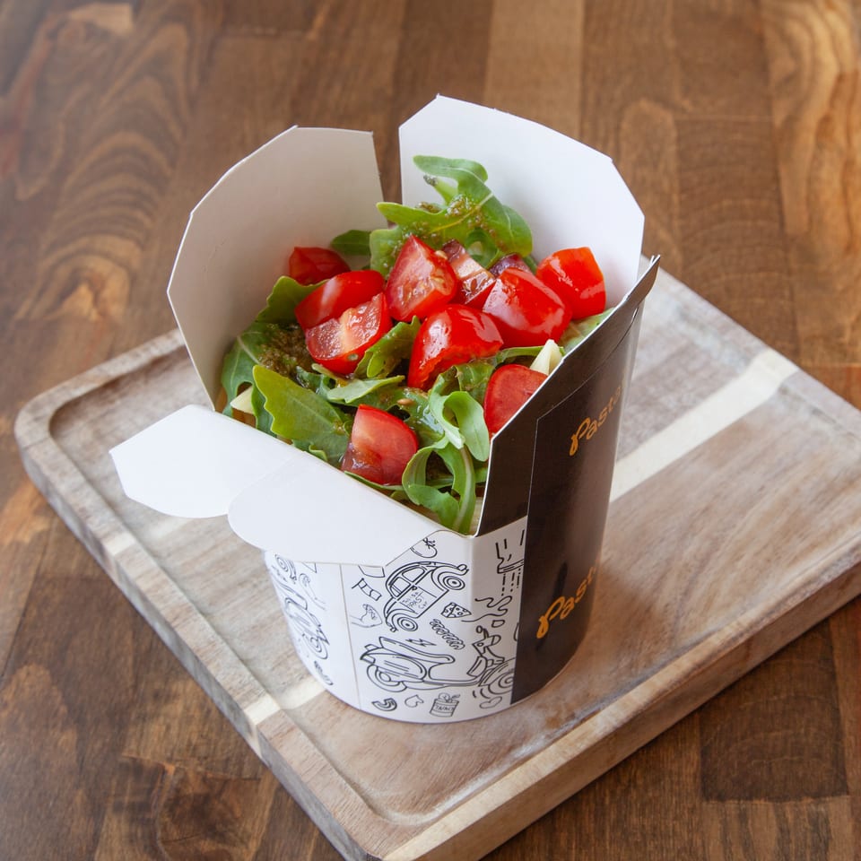 Fresh mixed green salad topped with chopped tomatoes in a branded takeaway cup on a wooden serving board.