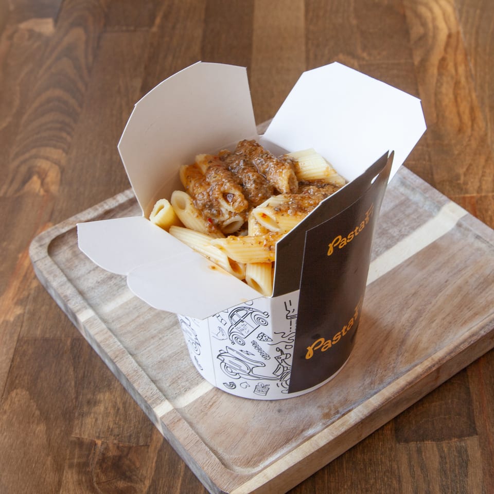 Takeout box of penne pasta topped with grilled chicken and creamy sauce on a wooden serving board.