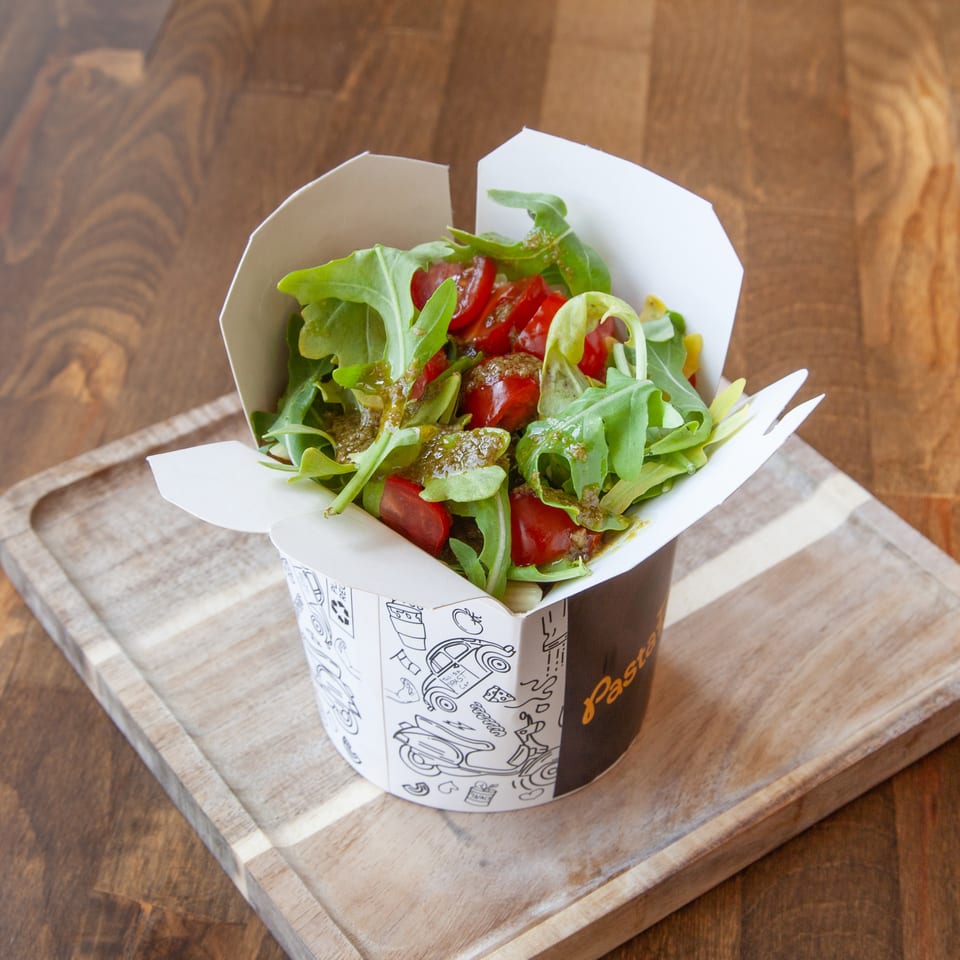 Fresh arugula and tomato salad served in a patterned takeaway cup on a wooden serving board.