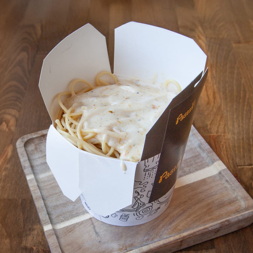 Spaghetti topped with creamy herb sauce in an open Pastamania takeaway box on a wooden serving board.