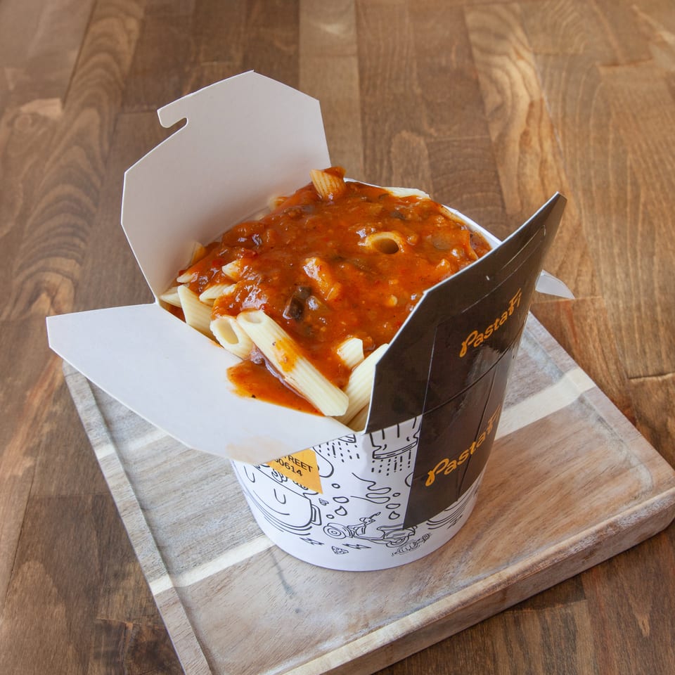 Penne pasta topped with chunky tomato sauce in a branded takeaway container on a wooden serving board.